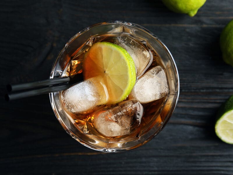 Glass of cocktail with cola, ice and cut lime on wooden background, top view